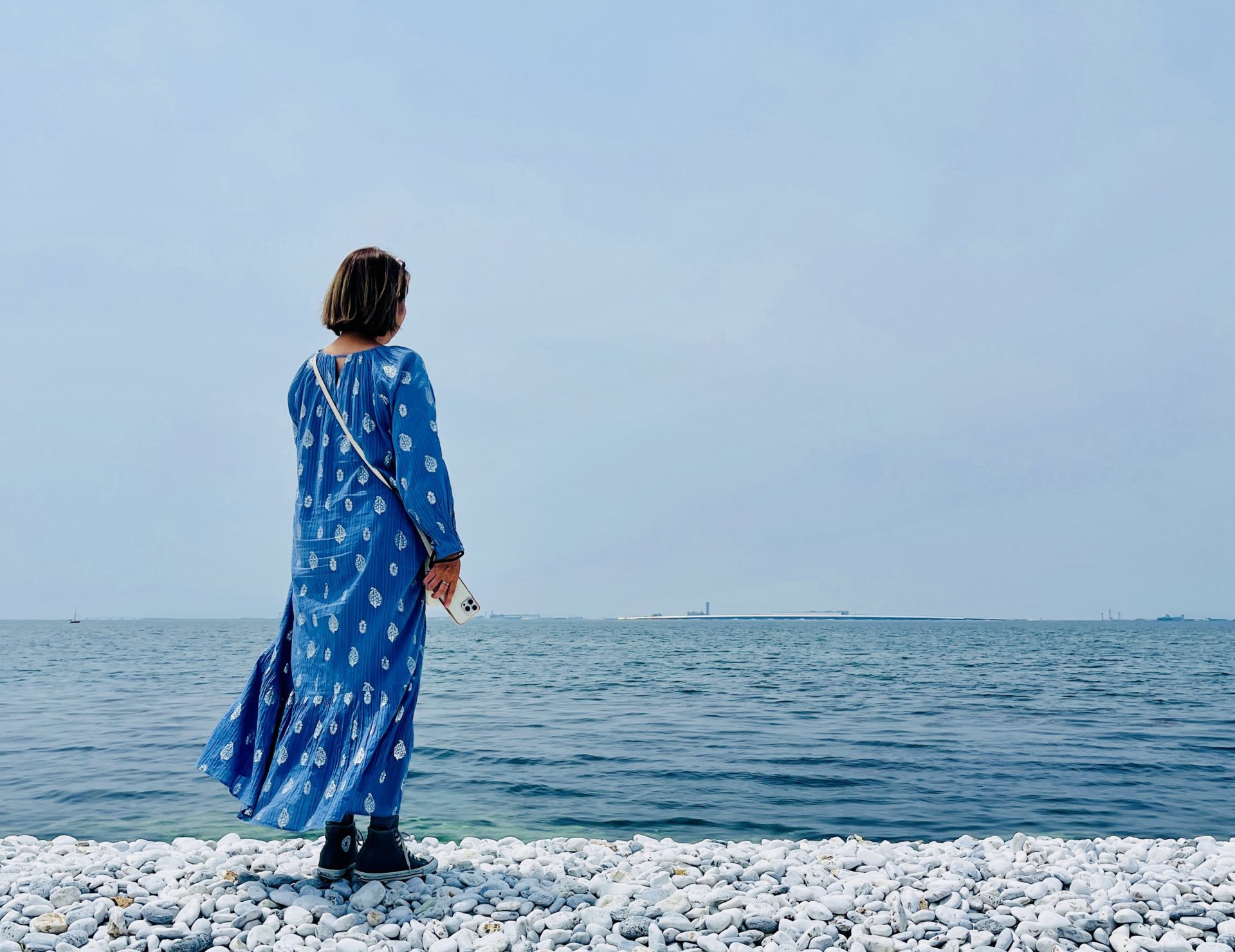 白い小石の海岸に立ち、青いワンピースの人物が海の水平線を見つめている
